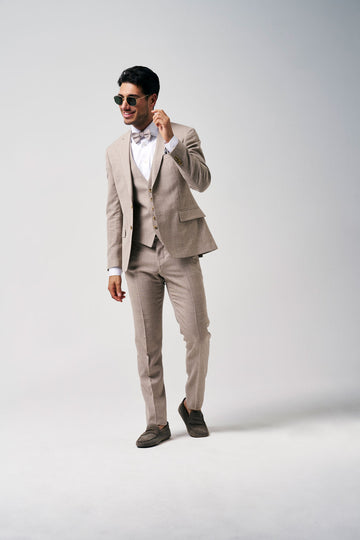 Mannelijk model in een beige driedelig kostuum met strik en zonnebril, wandelend tegen een lichte achtergrond.
