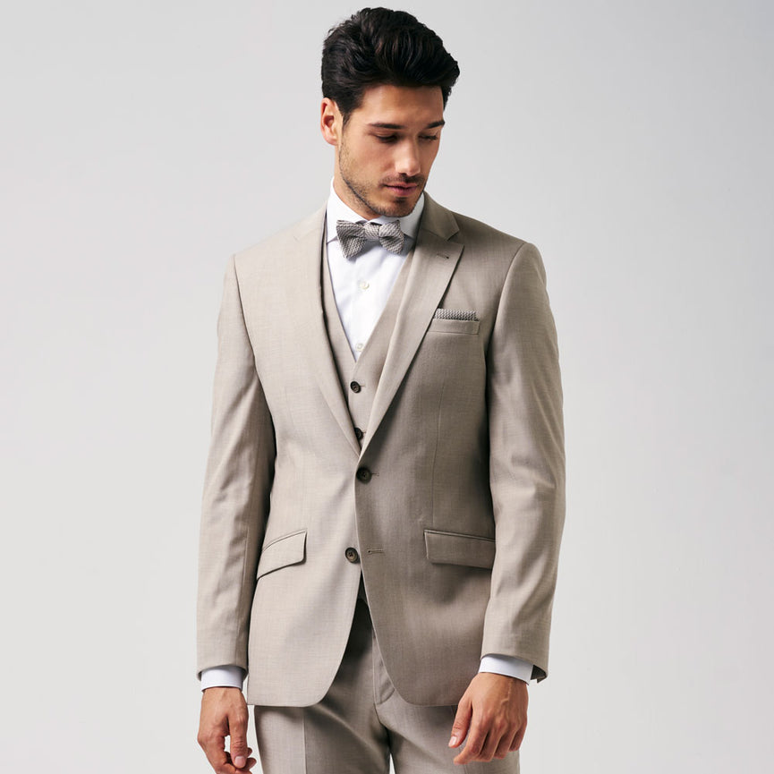 Mannelijk model in een beige driedelig kostuum met strik, gefotografeerd tegen een lichte achtergrond, kijkt naar beneden.